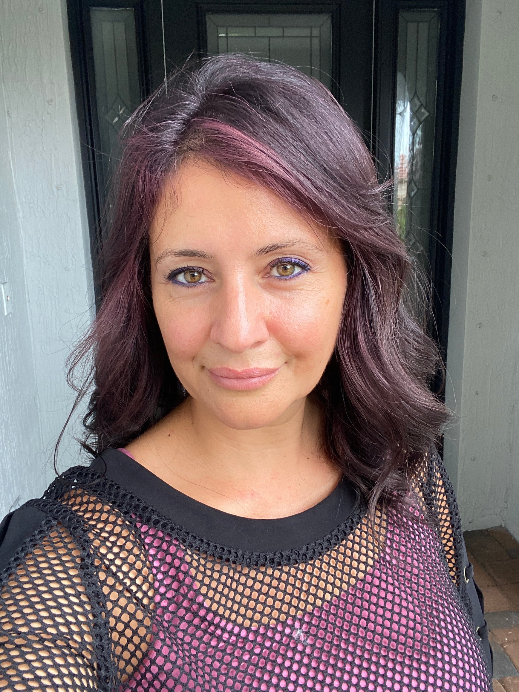 “Woman with shoulder-length wavy dark hair featuring subtle purple highlights, wearing a mesh top and smiling outdoors.”