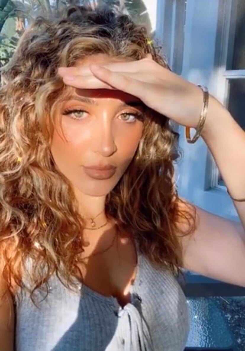 “Woman with sunlit curly blonde hair, wearing a gray top and shielding her face with her hand, posing outdoors in soft natural light.”