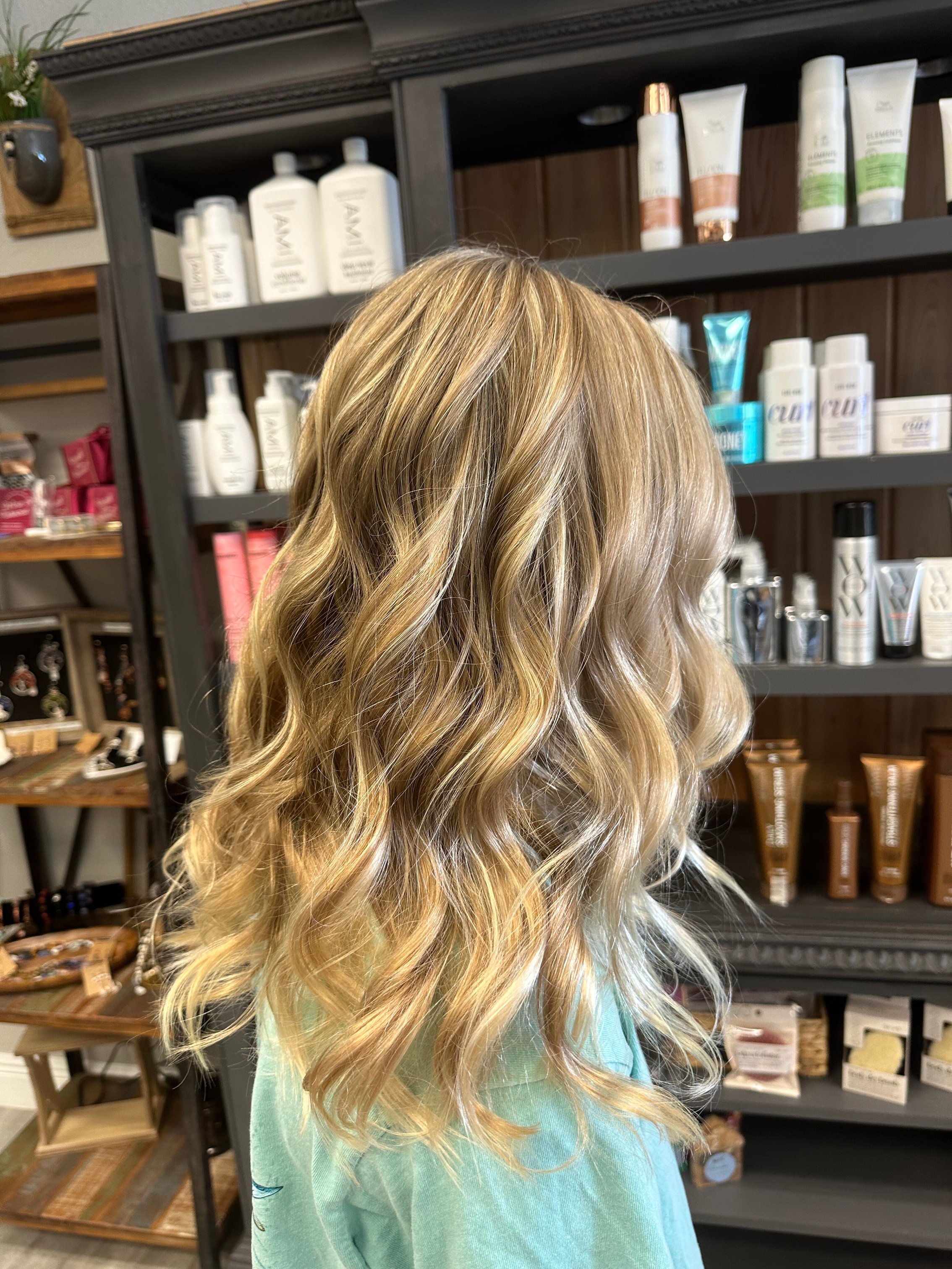 “Blonde hair styled into soft, flowing waves with a natural shine, displayed in a salon with shelves of hair products in the background.”
